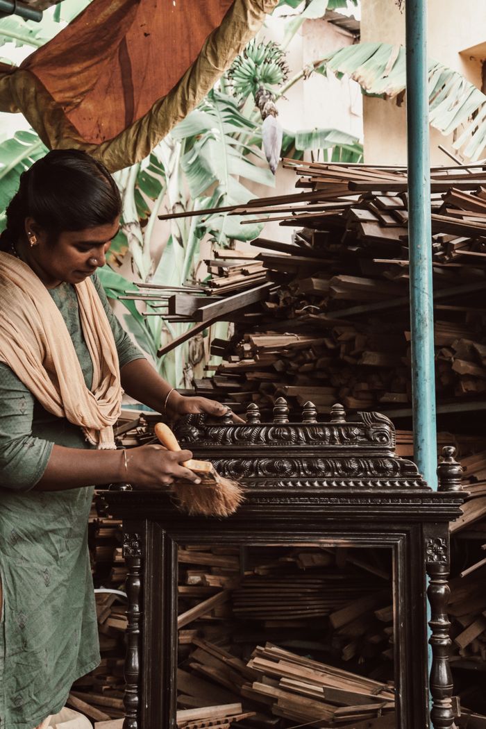 services-04 A woman in a green dress dusts elaborate wooden furniture in an artisan workshop surrounded by wooden planks.