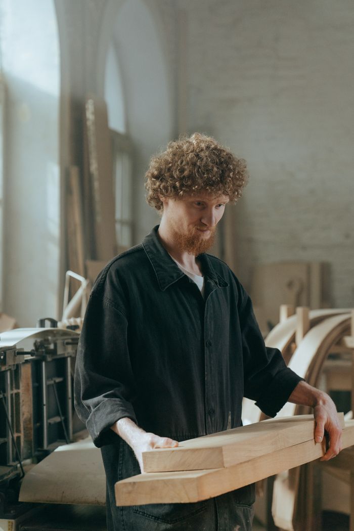 gallery-3 A bearded craftsman in a woodworking workshop holding timber planks.